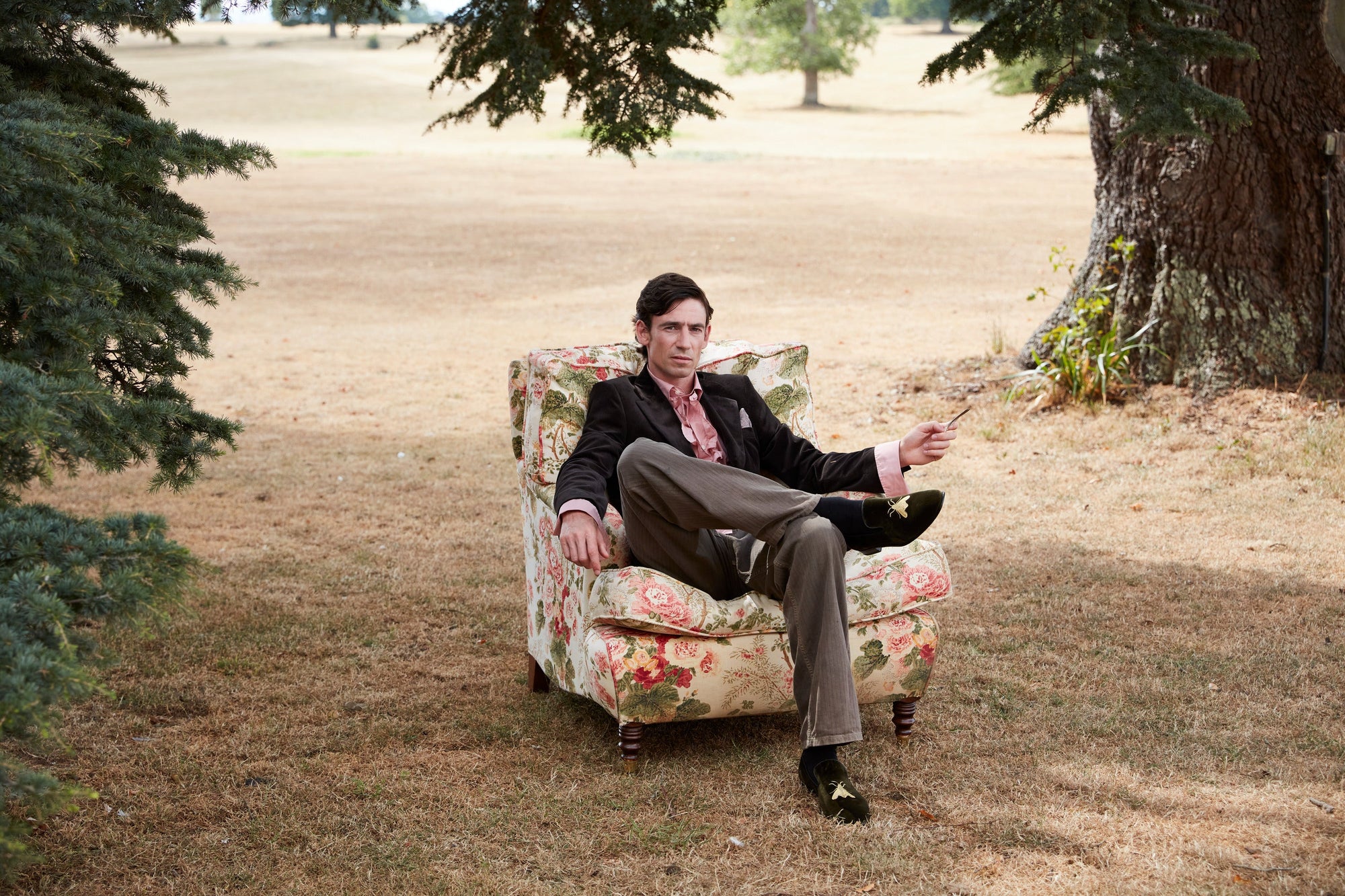 A chintz armchair carried onto the lawn at Neston Park beneath a cedar tree, with a model seated in a brown corduroy jacket and velvet slippers