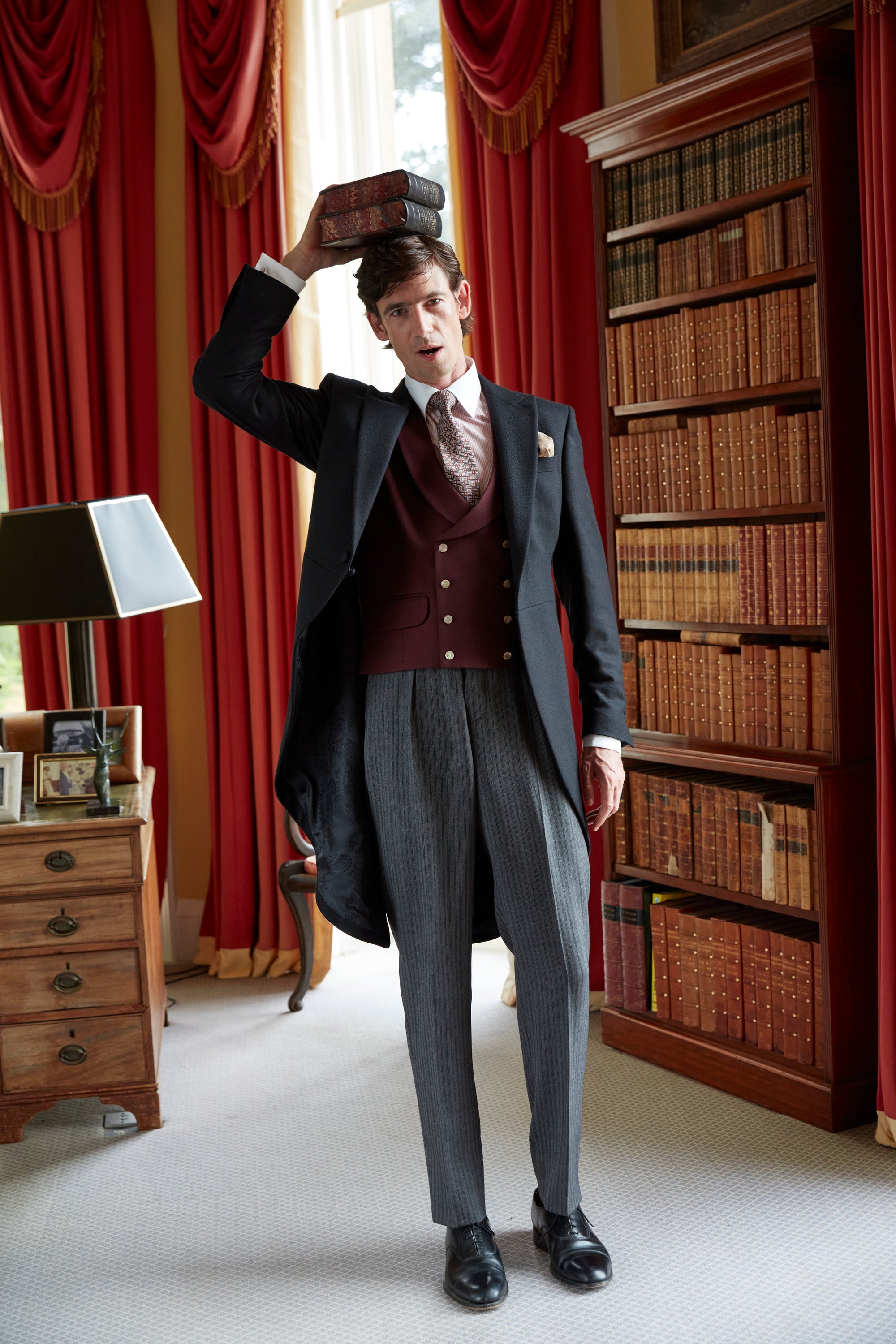 A model in a black morning coat and burgundy waistcoat reaching for a book in the library at Neston Park