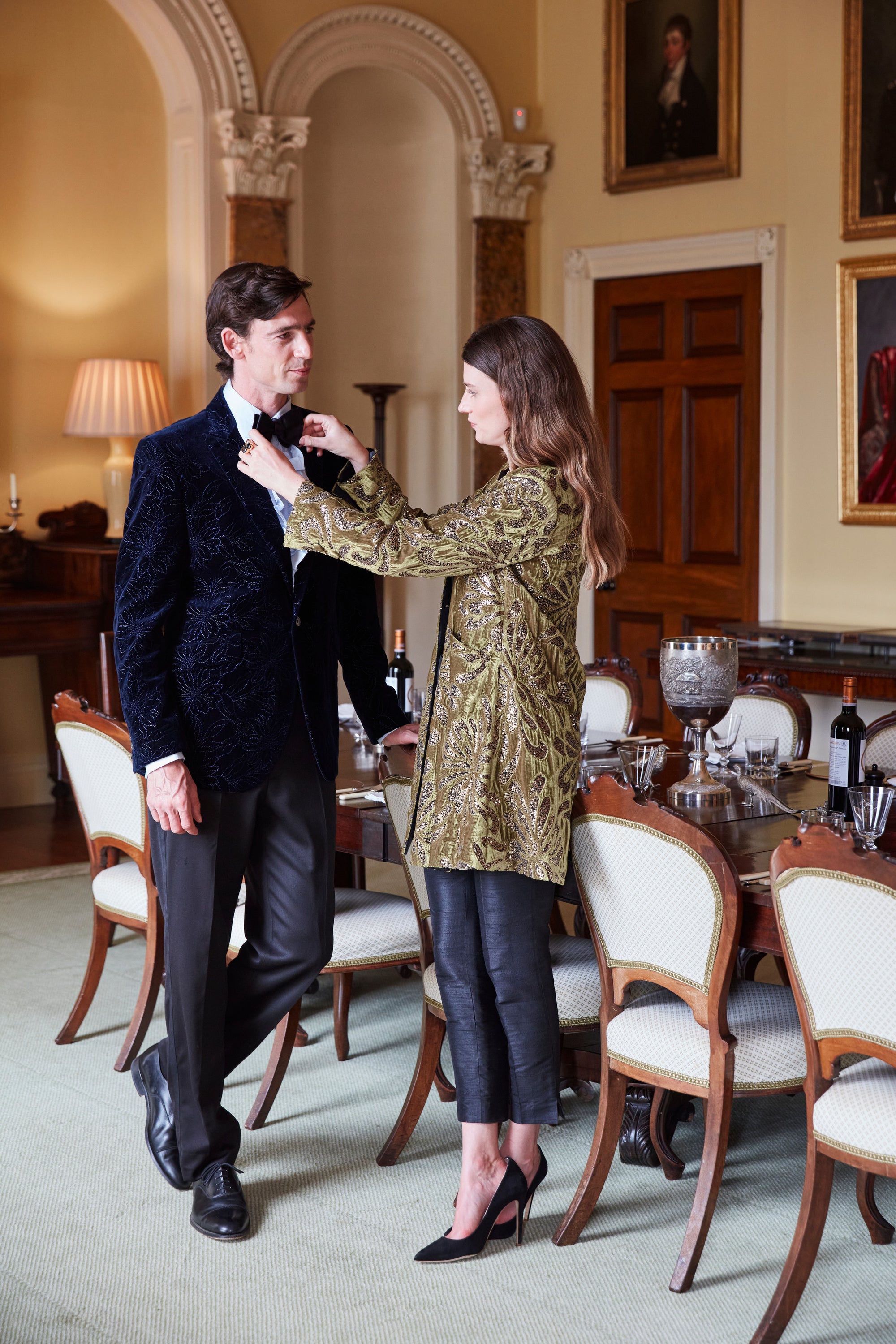 A couple dressed for the evening, she in a gold and olive brocade coat, he in a navy embroidered velvet dinner jacket