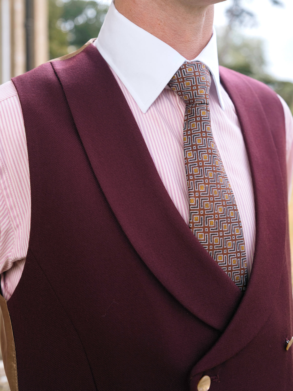 Close detail of a burgundy double breasted waistcoat, its shawl lapel, buttons and a woven silk tie