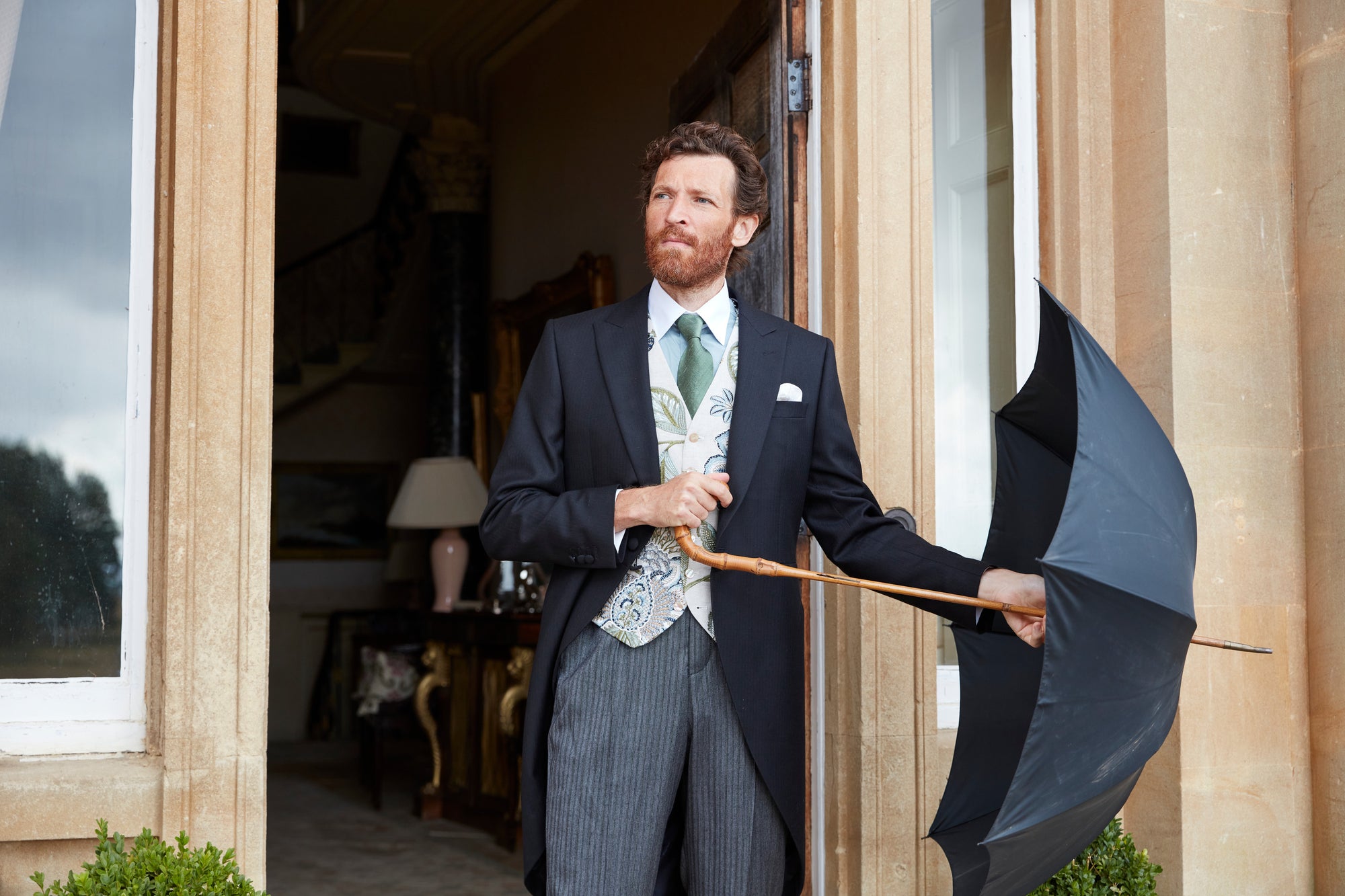 Favourbrook Autumn/Winter '26 menswear, photographed at Neston Park: a model in a morning coat with a patterned ivory waistcoat, holding a black umbrella at a window